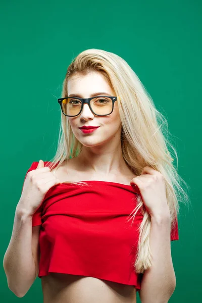 Portrait of Young Woman with Long Blond Hair, Eyeglasses and Bare Shoulders in Red Top Posing on Green Background in Studio. — Stock Photo, Image