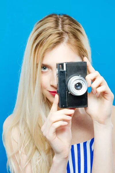 Linda mujer rubia en vestido de rayas de verano está fotografiando usando cámara retro sobre fondo azul en el estudio . — Foto de Stock