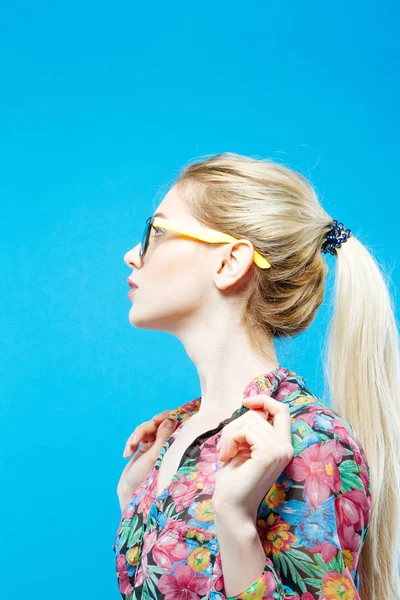 Vista laterale della ragazza carina con coda di cavallo lunga che indossa camicia colorata e occhiali su sfondo blu in studio . — Foto Stock