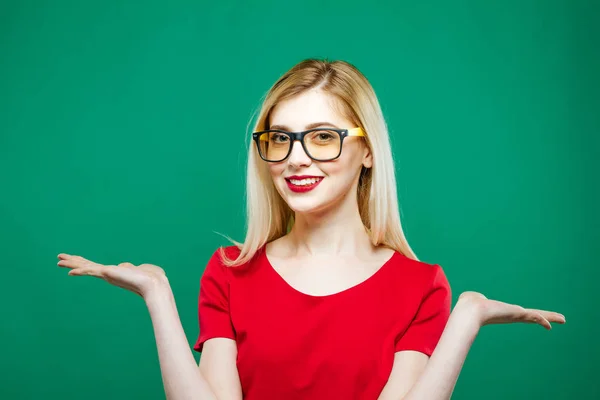 Ritratto di giovane bella donna con lunghi capelli biondi, occhiali da vista e top rosso che tiene lo spazio vuoto sulle sue due mani su sfondo verde in studio . — Foto Stock