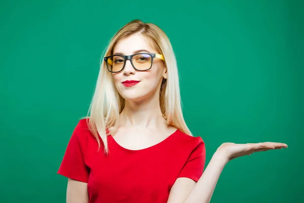 Ritratto di giovane bella donna con lunghi capelli biondi, occhiali da vista e top rosso che tiene lo spazio vuoto sulla sua mano su sfondo verde in studio . — Foto Stock