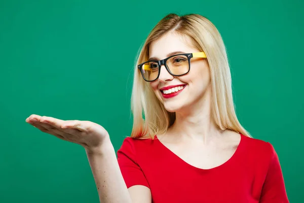 Donna sorridente con lunghi capelli biondi, occhiali da vista e top rosso che tiene lo spazio vuoto sulla mano su sfondo verde in studio . — Foto Stock