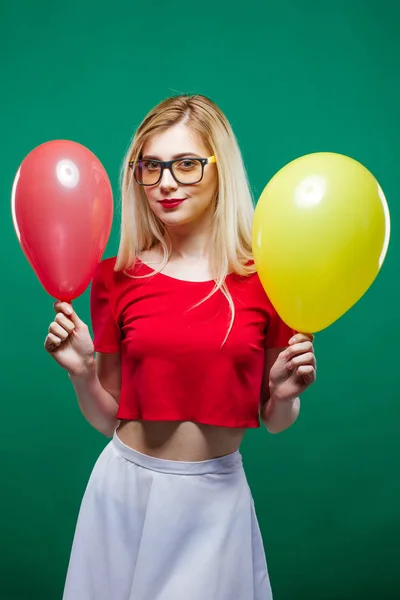 Ragazza del partito in occhiali alla moda è smorfia e guardando la fotocamera che tiene palloncini d'aria gialli e rossi nelle mani. Sorridente giovane donna divertirsi durante la celebrazione su sfondo verde . — Foto Stock