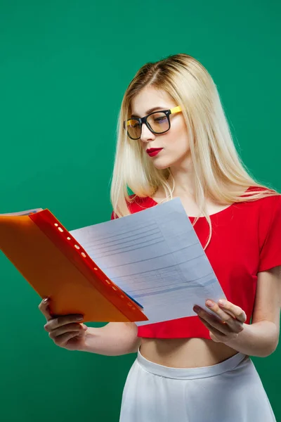 Bella studentessa in occhiali da vista, top corto rosso e gonna bianca sta leggendo qualcosa su sfondo verde in studio . — Foto Stock