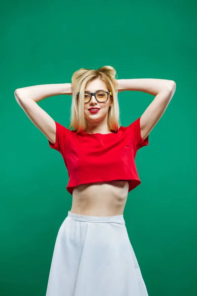Incredibile modello biondo con capelli lunghi e occhiali che indossano gonna bianca e breve top rosso è in posa in studio su sfondo verde . — Foto Stock
