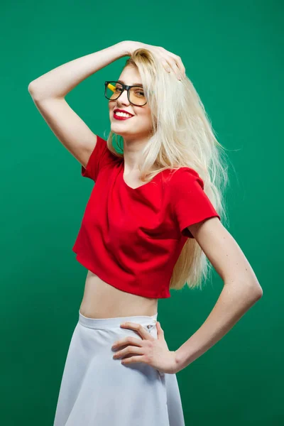Модная модель блондинки с длинным каблуком и очками, белой юбкой и красным топом появилась в студии на зеленом фоне . — стоковое фото