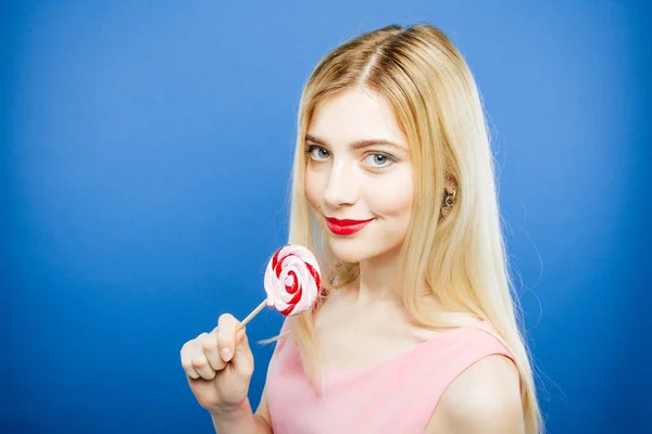 Pretty Blonde with Red Sensual Lips and Wonderful Smile is holding Sweet Lollipop near Her Face. Портрет удивительной девушки в розовом платье на голубом фоне в студии . — стоковое фото
