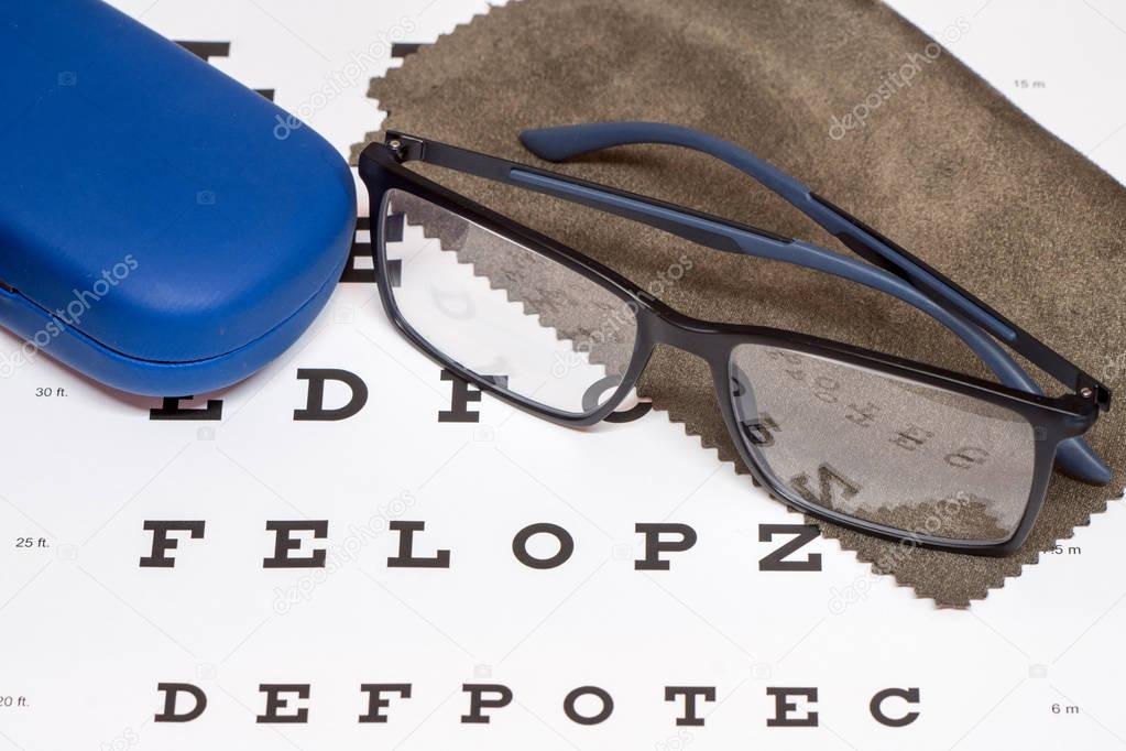 Reading black eyeglasses, brown microfiber cleaning cloth and blue protective case on white eye chart 