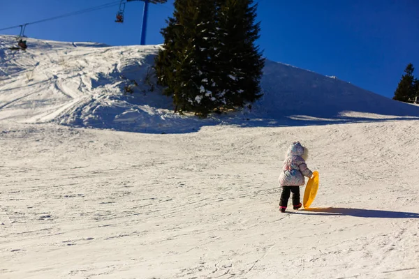 Ein kleines Mädchen in Winterkleidung schiebt einen Holzschlitten hinauf auf den schneebedeckten Hügel. aus niedriger Perspektive gesehen. — Stockfoto