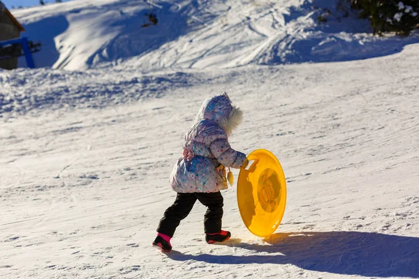 Ein kleines Mädchen in Winterkleidung schiebt einen Holzschlitten hinauf auf den schneebedeckten Hügel. aus niedriger Perspektive gesehen. — Stockfoto