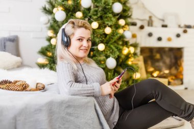 Happy woman enjoying her music at Christmas relaxing on a sofa in front of the tree with a beaming smile clasping her mobile to her chest.