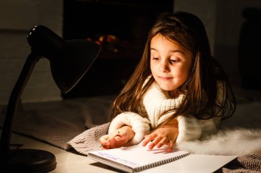 Küçük, güzel bir kız. Lambanın altına deftere yazıyor.