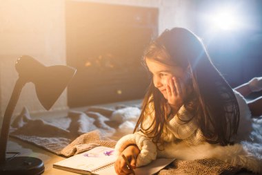 Küçük, güzel bir kız. Lambanın altına deftere yazıyor.