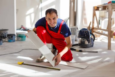 Üstad iş kıyafetleri giyiyor, fayans döşemeyi kontrol ediyor, bina seviyesini kullanıyor, ayarlamalar yapıyor, profesyonel onarım işi yapıyor. 