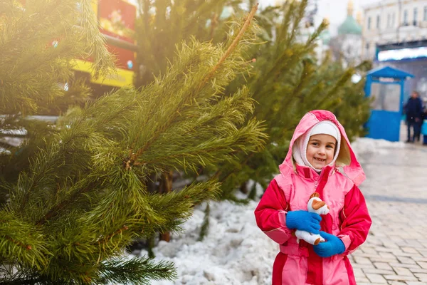可爱的小女孩带着圣诞树笑着 穿着冬衣和玩具的快乐孩子在圣诞市场上选择圣诞树 背景上点着灯 庆祝概念 — 图库照片