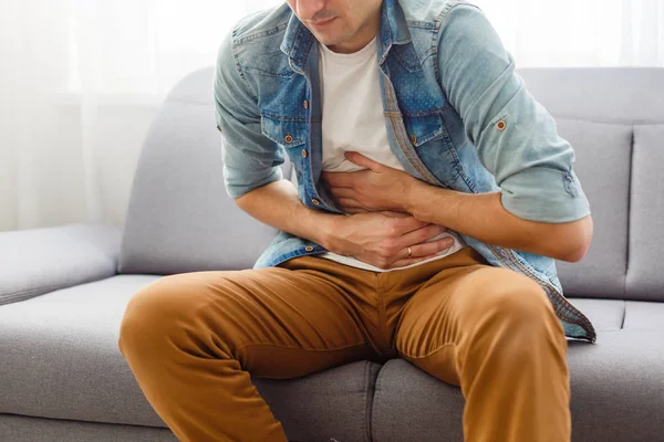 Manden sidder på en grå sofa og holder maven. Medicin - Stock-foto