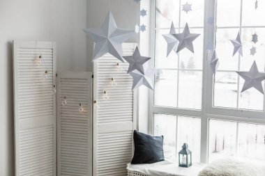 Büyük, parlak bir pencere. Pencere pervazında gri örülü ekose kumaş, hediyeler ve parlayan otlaklar var. Pencerenin arka planında kağıttan Noel yıldızları asılı..