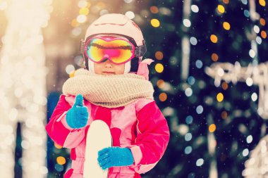 Kış tatilinde gözlük ve kask takan küçük bir kız kameranın arkasında kayak tutuyor. Gülümsüyor, heyecanlı, yakın plan bulanıklığı çekiyor.