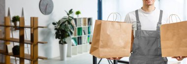 Elinde kese kâğıdı olan genç adam, yakın plan. Tişört giyiyor, açık gri arka plan. Yemek teslimatı.