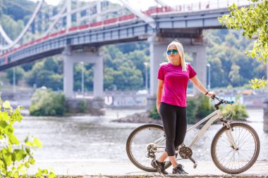 Köprü arka planında bisiklete binen zayıf genç bir kadının arka görüntüsü. Aktif yaşam tarzı ve tatil kavramı.
