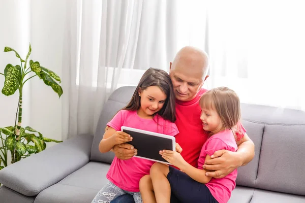 Un homme âgé et une petite fille assis sur ses genoux sont assis dans une pièce. Grand-père tenant une tablette dans sa main et regardant sa petite-fille — Photo