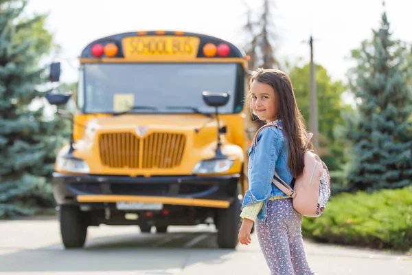 Kislány áll egy nagy iskolabusz ajtó mellett a rózsaszín hátizsákjával.. — Stock Fotó