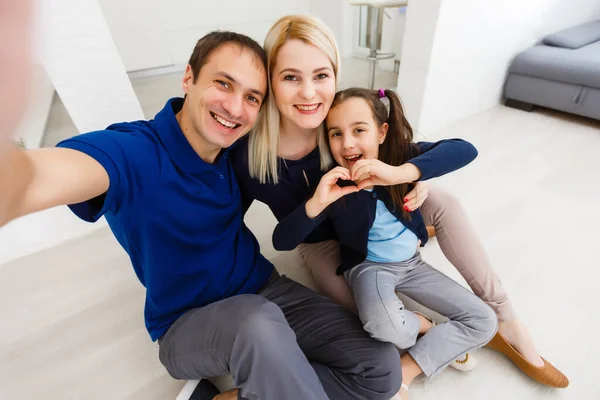 Όμορφη νεαρή μητέρα, ο πατέρας και η κόρη τους βγάζουν selfie χρησιμοποιώντας ένα τηλέφωνο και χαμογελώντας ενώ κάθονται στο σπίτι — Φωτογραφία Αρχείου