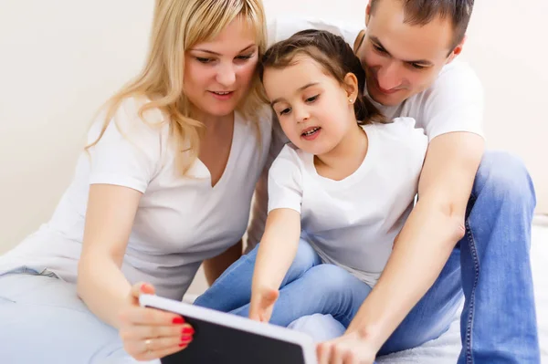 Una atractiva familia feliz, de madre, padre e hija sentados en un sofá en casa divirtiéndose con una tableta — Foto de Stock