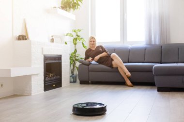 Kadın kanepede dinlenirken robot süpürgesi odayı temizliyor.