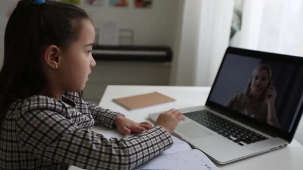 Smart Little Girl hace la tarea en su sala de estar. Ella se sienta en su escritorio, utiliza el ordenador portátil y escribe con una pluma en sus libros de texto. — Vídeo de stock