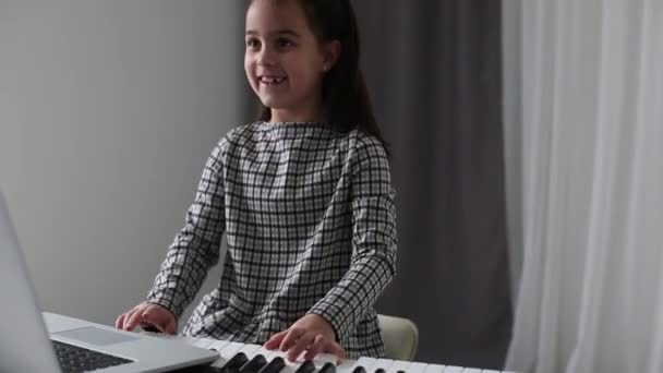 Маленька дівчинка дивиться музичний урок онлайн і вчиться вдома. маленька дівчинка практикує свої навички піаніно, дивлячись на екран комп'ютера після професора на відеодзвінок . — стокове відео