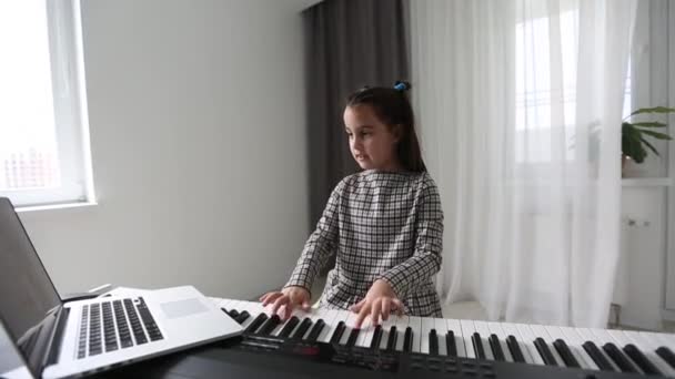 Маленькая девочка смотрит уроки музыки в Интернете и учится дома. маленькая девочка отрабатывает свои навыки игры на фортепиано, глядя на экран компьютера вслед за профессором по видеосвязи. — стоковое видео