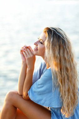 Gün batımında deniz kenarında oturup rahatlayan genç sarışın bir kadının portresi.