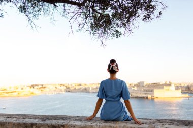 İnce, esmer, genç bir kadın denizin ve şehrin arka planına karşı yüksekte oturur ve doğayı düşünür. Kadın siluetine odaklan. Arka plan.