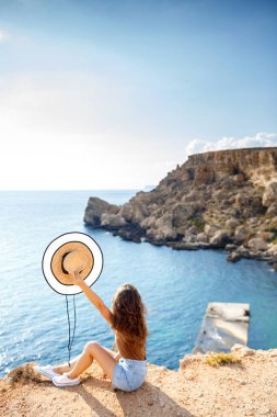 Şapkalı ince bir kadın turist denizin arka planında bir kayanın üzerinde oturur ve doğayı düşünür. Arka plan