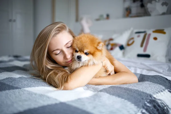 Mutlu sarışın kadın köpeği Spitz ile yatakta uzanıyor. Bir kadın ve bir köpeğin portresi..