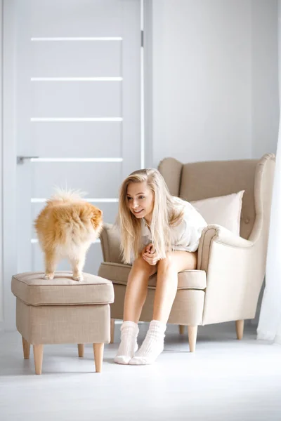 Happy blonde woman is sitting on a chair near her dog. Dog breed Pomeranian.
