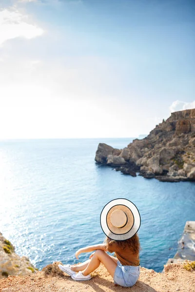 Şapkalı ince bir kadın turist denizin arka planında bir kayanın üzerinde oturur ve doğayı düşünür. Arka plan