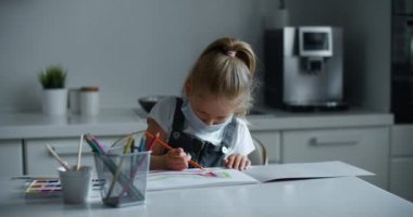 4-7 yaşlarındaki bir kız çocuğu koruyucu bir maskeyle evde oturuyor ve kağıda resim yapıyor. Çocuk resme odaklanmış bir gökkuşağı çiziyor. Büyük kadın portresi. Coronavirus ve izolasyon kavramı
