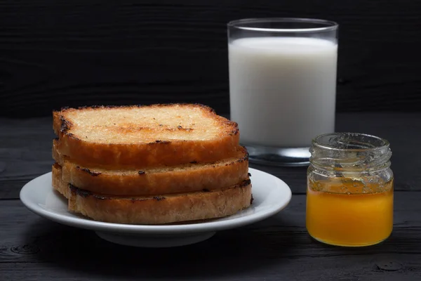 Tostar en un plato con leche y una lata de miel. Sobre un fondo de madera negro — Foto de Stock