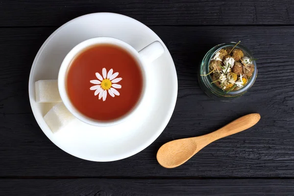 Té con cubos de azúcar, una cuchara de madera y manzanilla en un frasco sobre un fondo negro — Foto de Stock