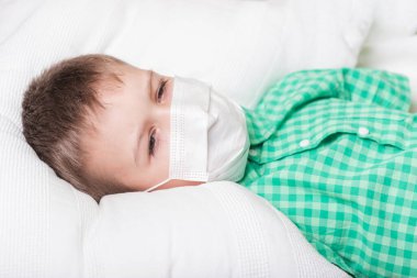 Tıbbi maskeli çocuk yatakta yatıyor.