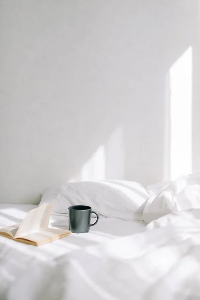 Yatağın üzerindeki parlak yatak odasında bir fincan kahve ya da çayın yanında açık eski bir kitap var. Güneş ışınlarının arka planına karşı. Dikey fotoğraf