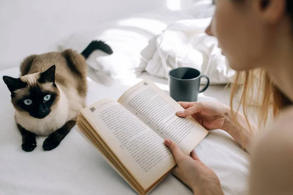 Young girl reads a book in bed in a bright bedroom next to a cat and a cup of coffee
