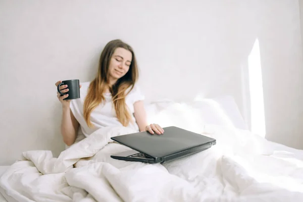Young beautiful girl starts to work on a laptop in bed holding a cup of coffee in her hand