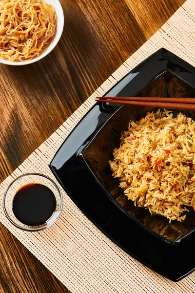 Siyah kare tabak, yemek çubukları, soya sosu, koyu ahşap masa ve malzemeler sebzeli pilav
