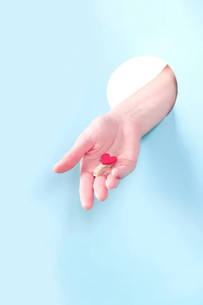 Крупный план человеческой руки, выступающей через отверстие в синем фоне, держа красное сердце . — стоковое фото