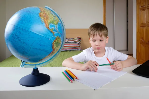 Distance learning, geography lesson. The boy is sitting on the bed with a globe at the laptop. Social distancing and self isolation in quarantine lockdown.