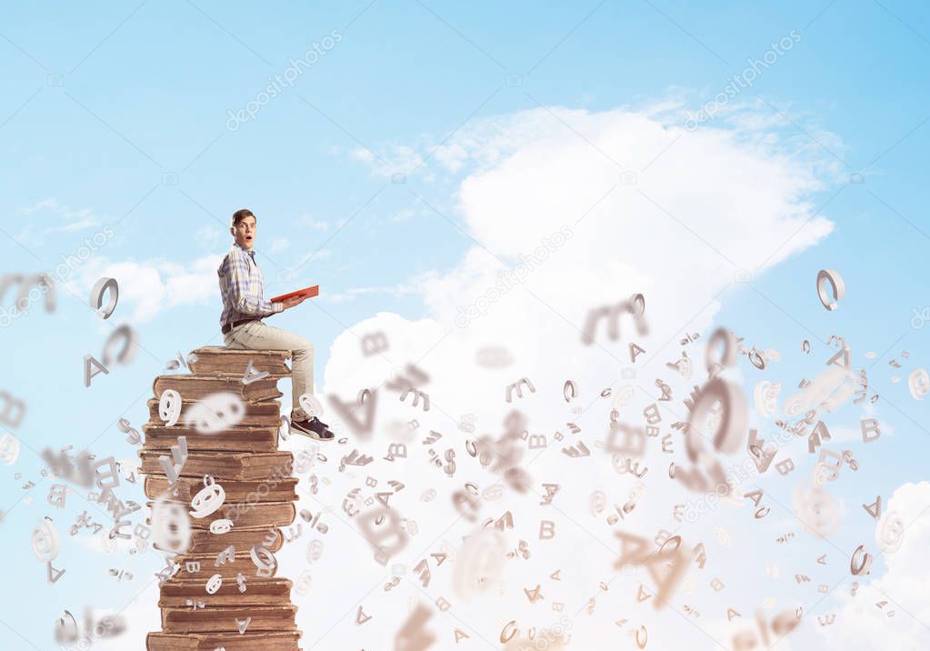 Young businessman holding book while sitting on pile of books and symbols flying around