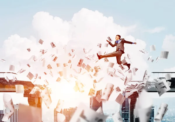 Businessman jumping over gap with flying paper documents in concrete bridge as symbol of overcoming challenges with sunlight skyscape on background